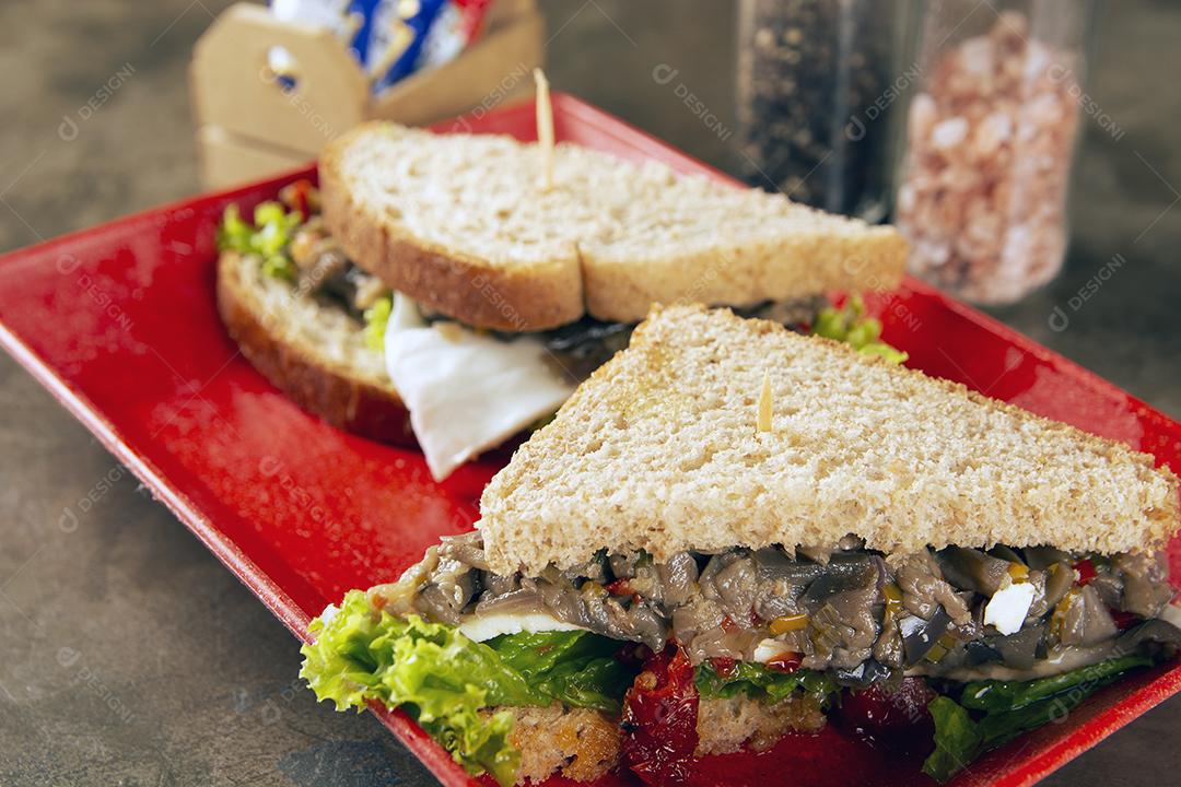 Sanduíche de berinjela, alface, tomate e pão simples em um prato vermelho sobre fundo de pedra.