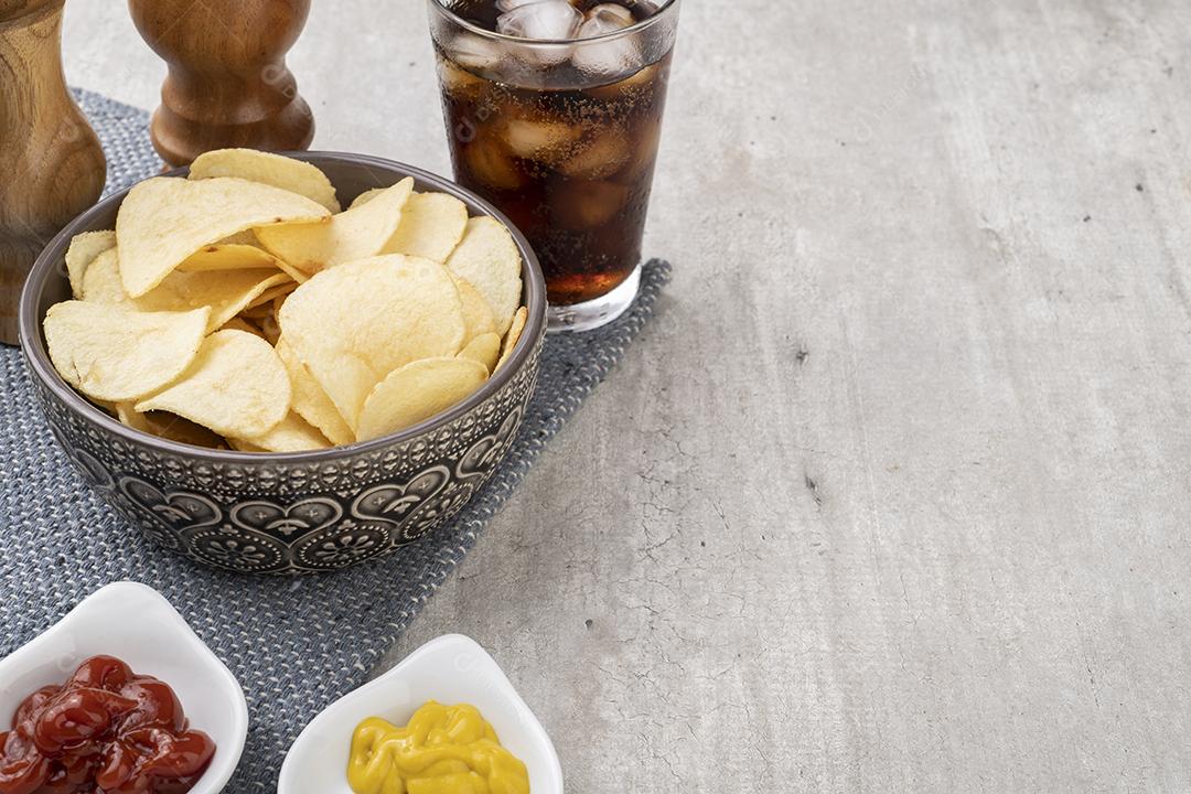 Batata frita em uma tigela, pimenta, sal, mostarda, ketchup e um copo de refrigerante. Fundo cinza claro