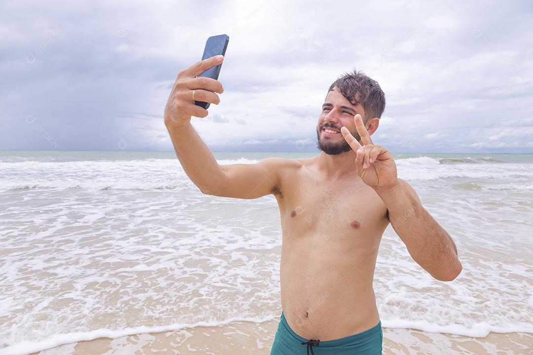 Homem bonito feliz fazendo uma selfie com smartphone na praia. Homem de férias na praia tirando foto com celular
