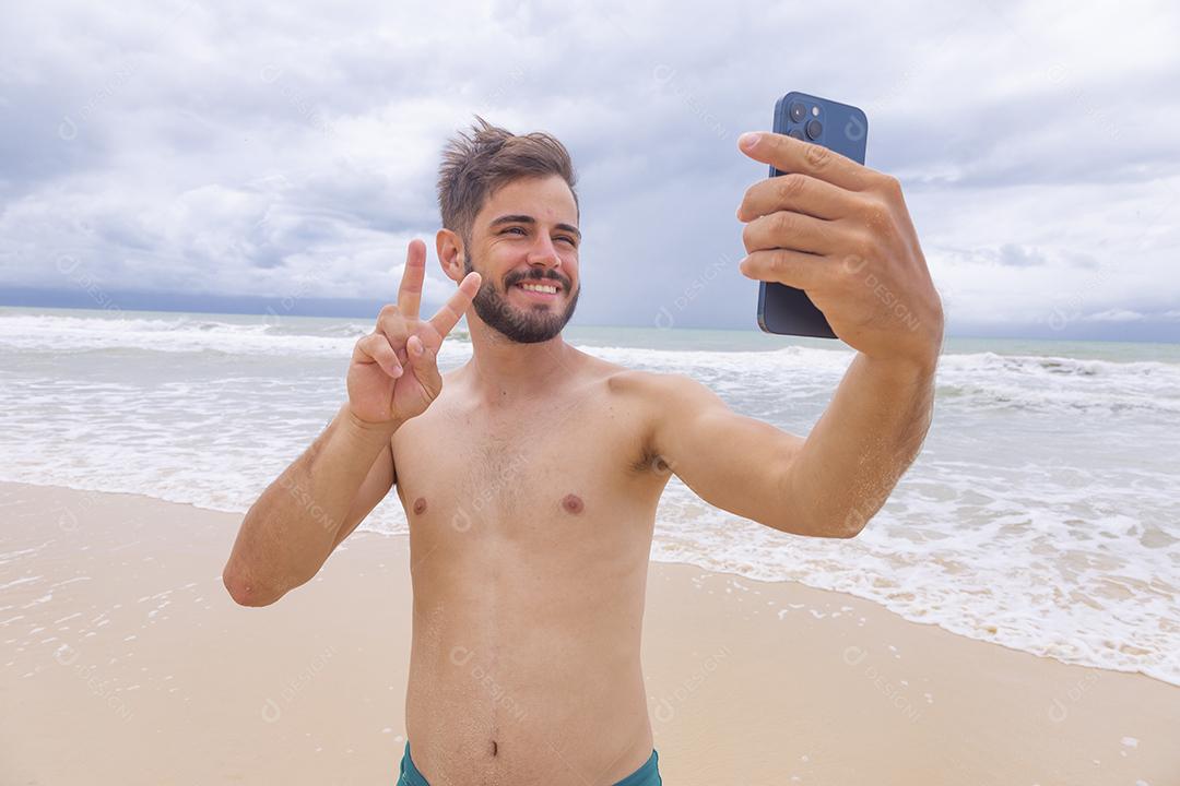 Homem bonito feliz fazendo uma selfie com smartphone na praia. Homem de férias na praia tirando foto com celular