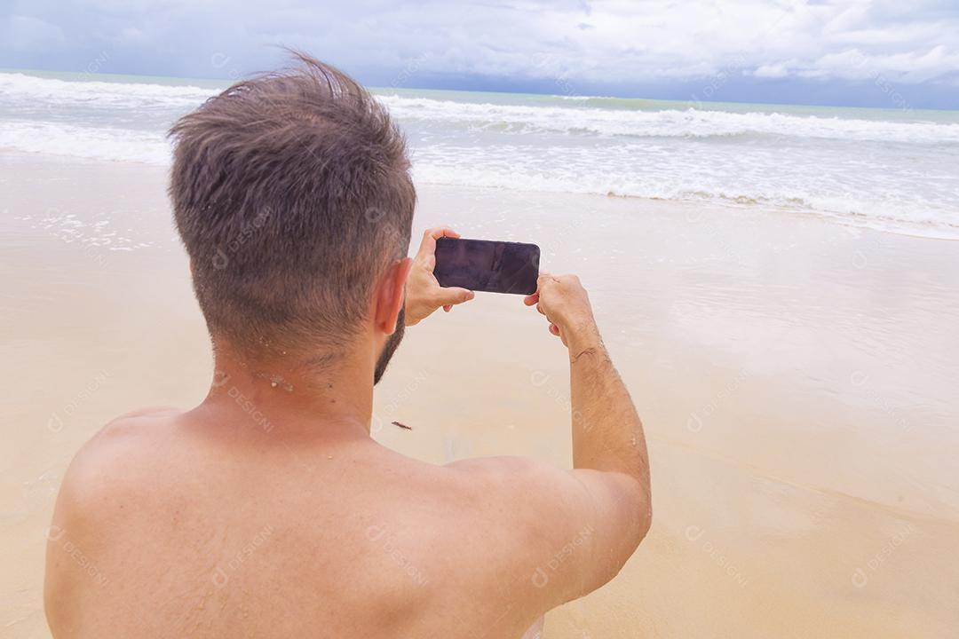 Rapaz olhando para a tela do celular em branco com espaço de cópia na areia da praia. Feriado