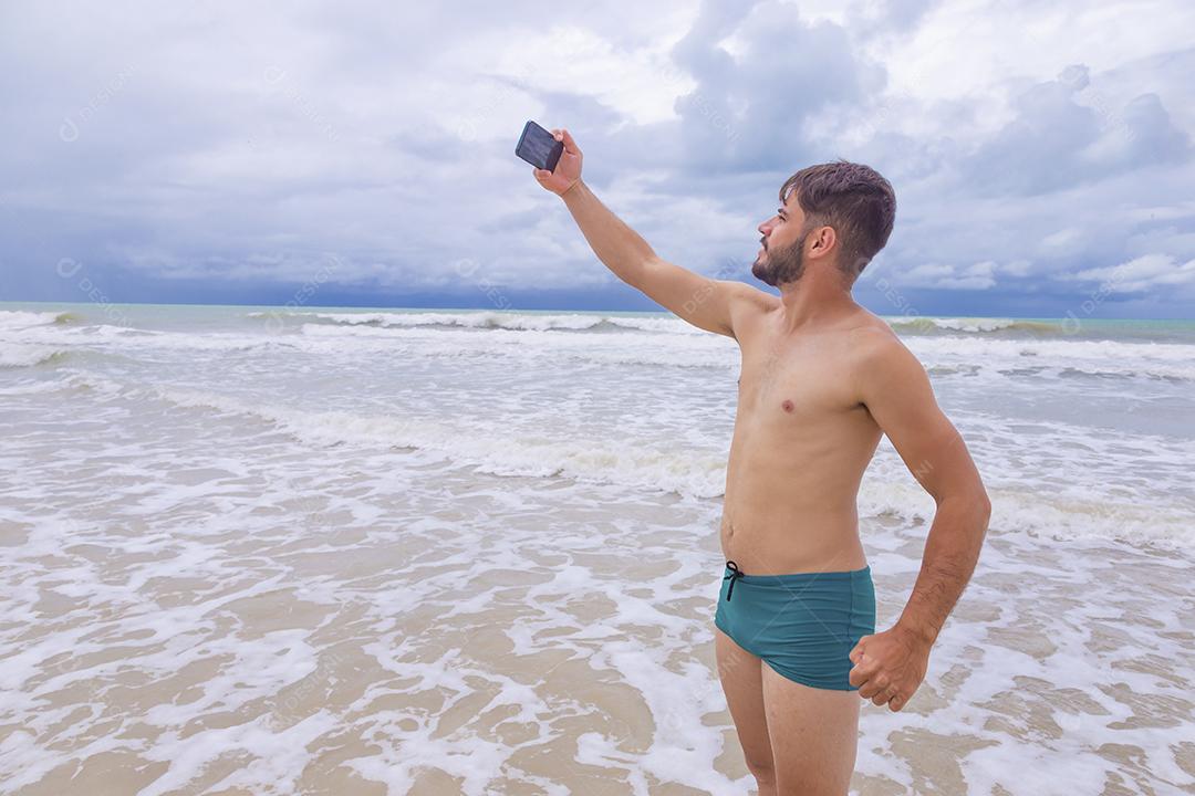 Homem bonito feliz fazendo uma selfie com smartphone na praia. Homem de férias na praia tirando foto com celular