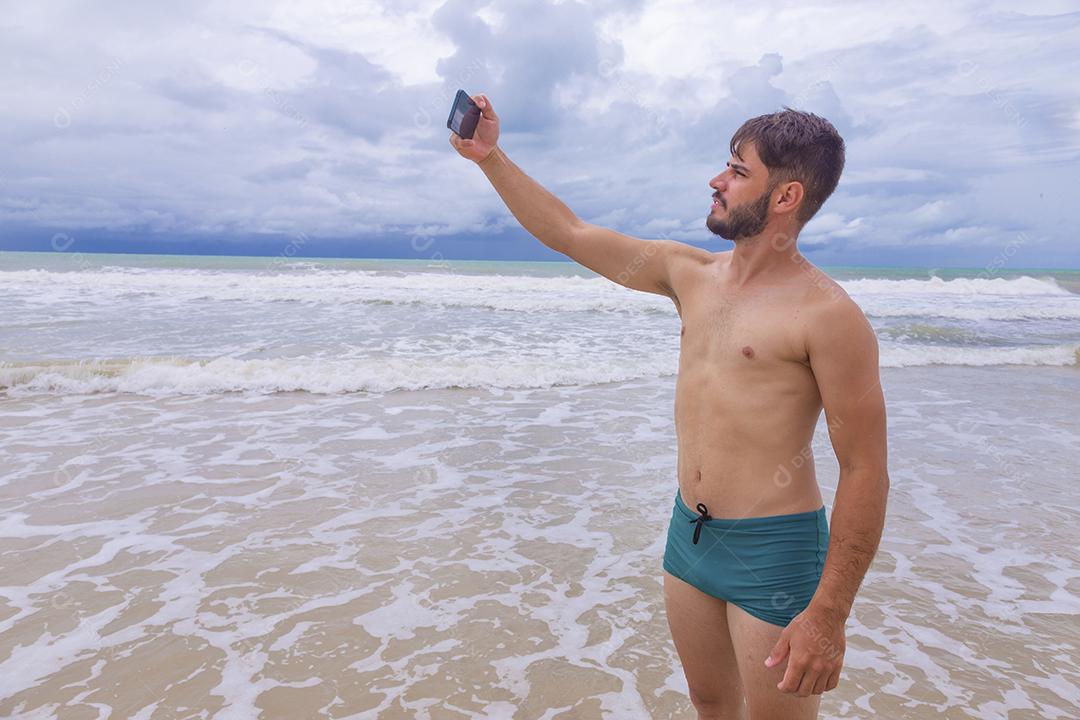 Homem bonito feliz fazendo uma selfie com smartphone na praia. Homem de férias na praia tirando foto com celular