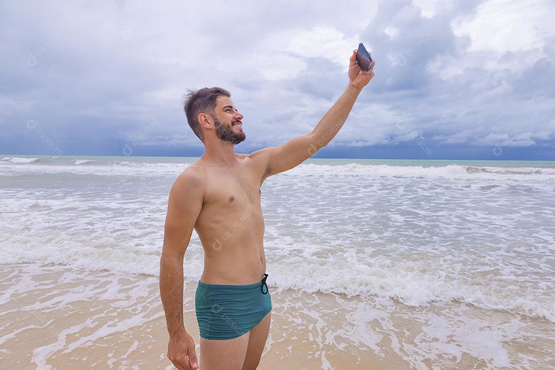 Homem bonito feliz fazendo uma selfie com smartphone na praia. Homem de férias na praia tirando foto com celular