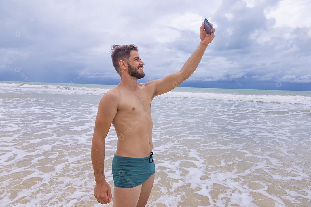 Homem bonito feliz fazendo uma selfie com smartphone na praia. Homem de férias na praia tirando foto com celular