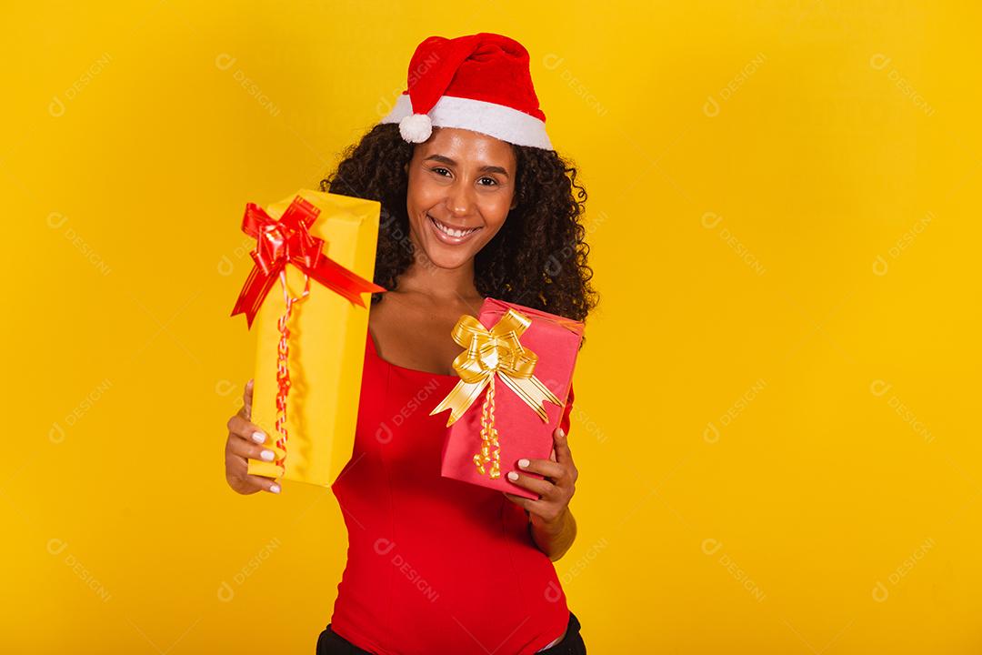 Linda mulher negra com chapéu de papai noel segurando o presente de feliz natal em fundo amarelo.