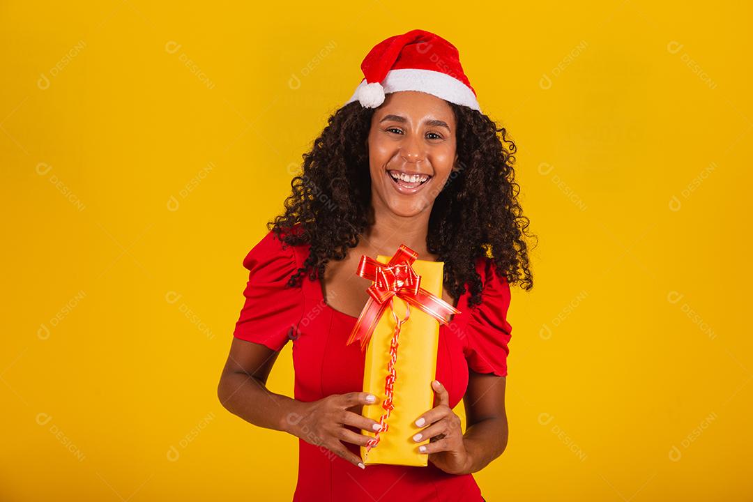 Linda mulher negra com chapéu de papai noel segurando o presente de feliz natal em fundo amarelo.