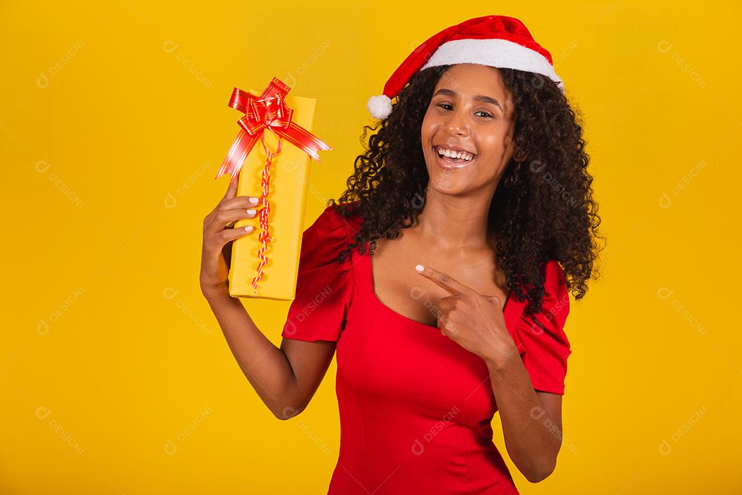 Linda mulher negra com chapéu de papai noel segurando o presente de feliz natal em fundo amarelo.
