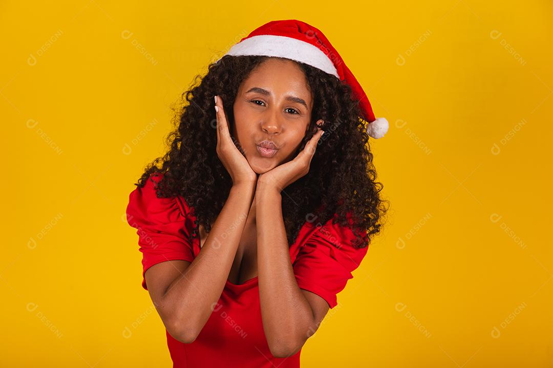 Mulher afro usando chapéu de papai noel franzindo os lábios fazendo beicinho ou mandando beijos em fundo amarelo