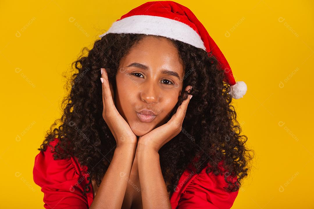 Mulher afro usando chapéu de papai noel franzindo os lábios fazendo beicinho ou mandando beijos em fundo amarelo