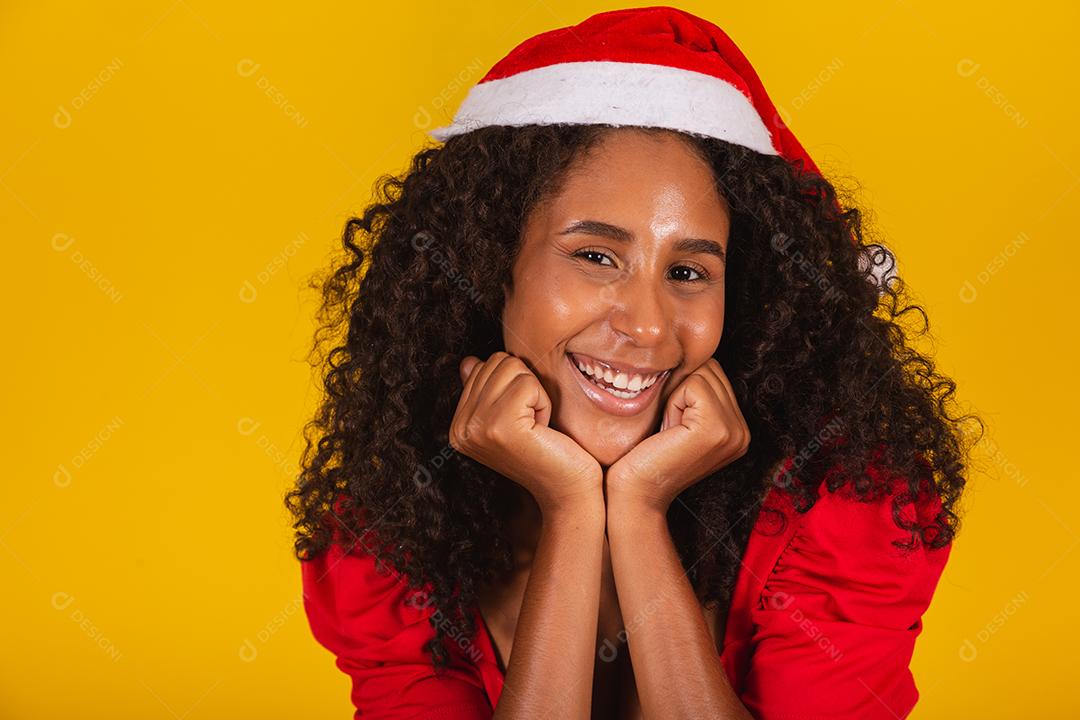 Mulher afro usando chapéu de papai noel franzindo os lábios fazendo beicinho ou mandando beijos em fundo amarelo