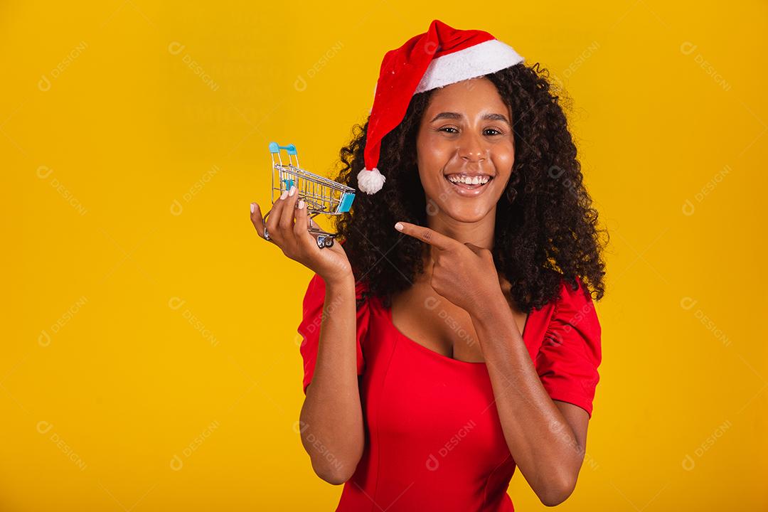 Mulher de chapéu de Papai Noel fazendo compras de Natal. Ela está mostrando um mini carrinho