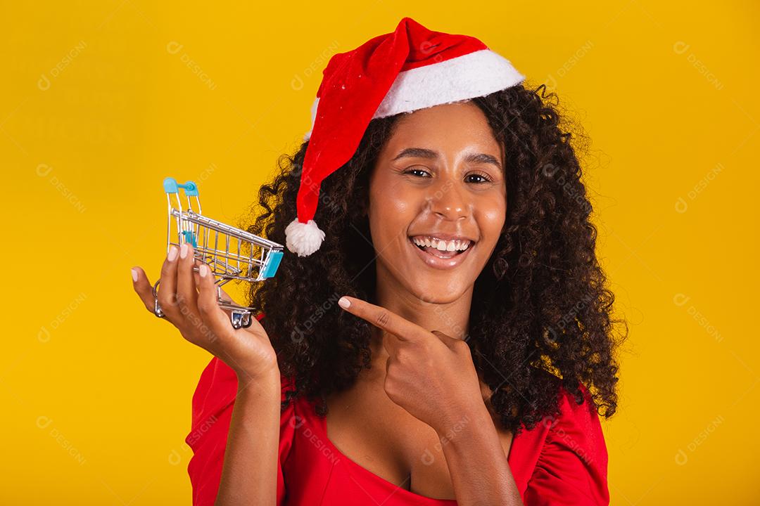 Mulher de chapéu de Papai Noel fazendo compras de Natal. Ela está mostrando um mini carrinho
