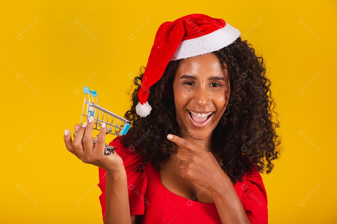 Mulher de chapéu de Papai Noel fazendo compras de Natal. Ela está mostrando um mini carrinho