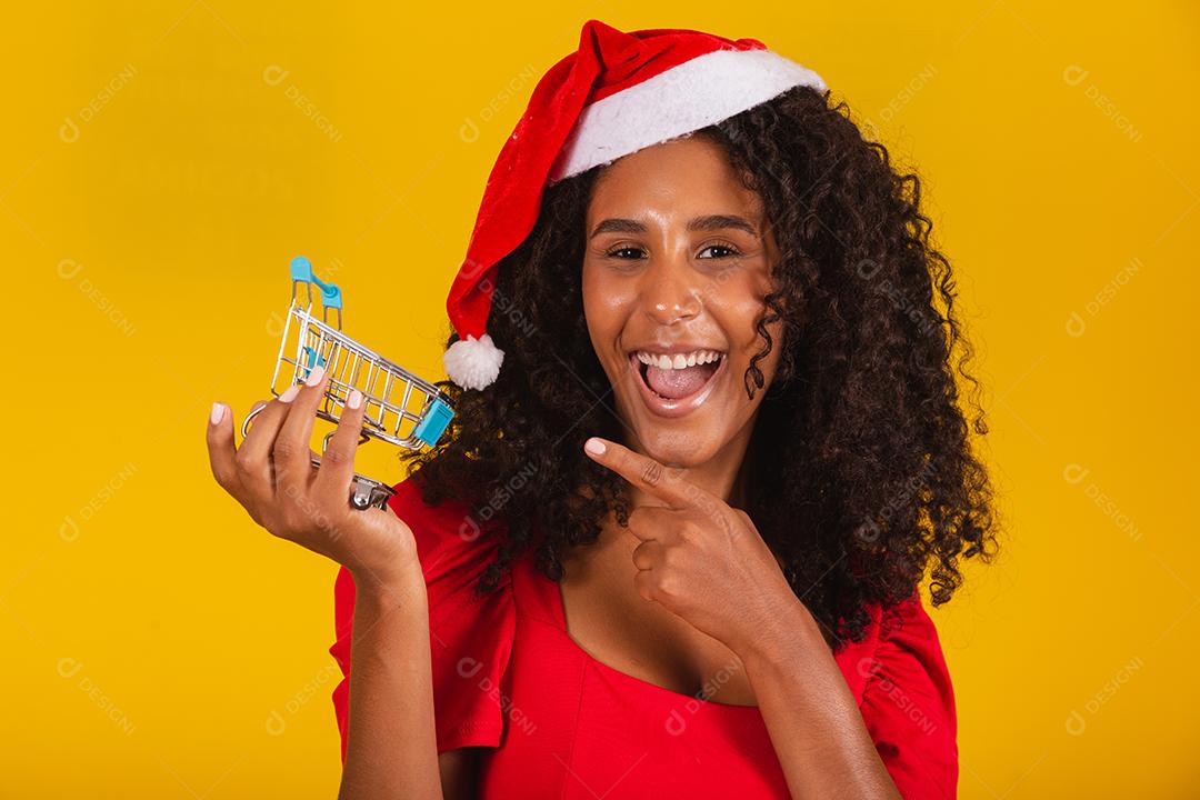 Mulher de chapéu de Papai Noel fazendo compras de Natal. Ela está mostrando um mini carrinho