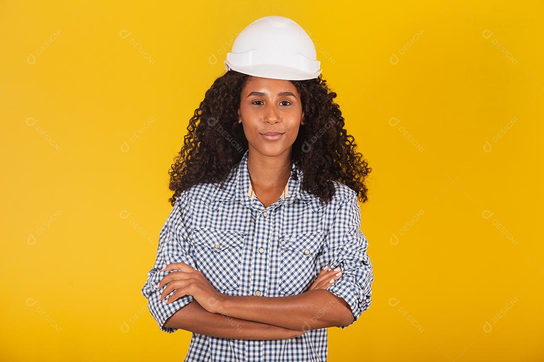 Engenheira ou arquiteta em fundo amarelo sorrindo com os braços cruzados.