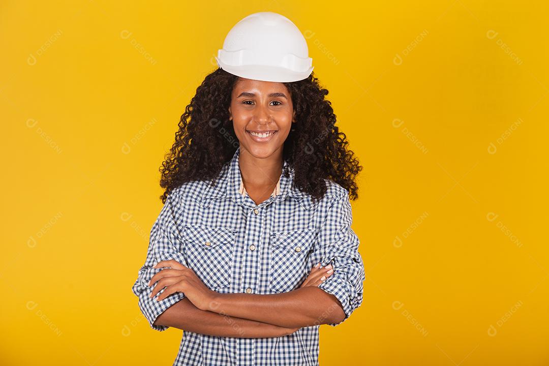 Engenheira ou arquiteta em fundo amarelo sorrindo com os braços cruzados.