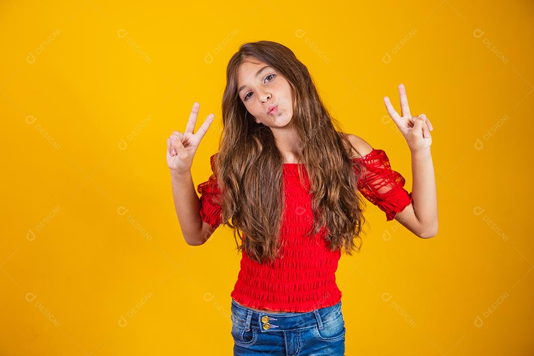 Menina pré-adolescente encantadora e delicada fazendo um sinal de V sorrindo isolado em fundo amarelo.