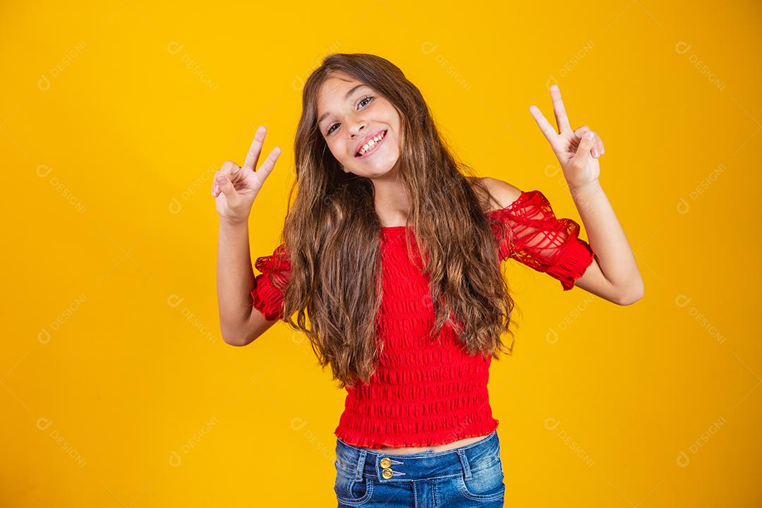 Menina pré-adolescente encantadora e delicada fazendo um sinal de V sorrindo isolado em fundo amarelo.