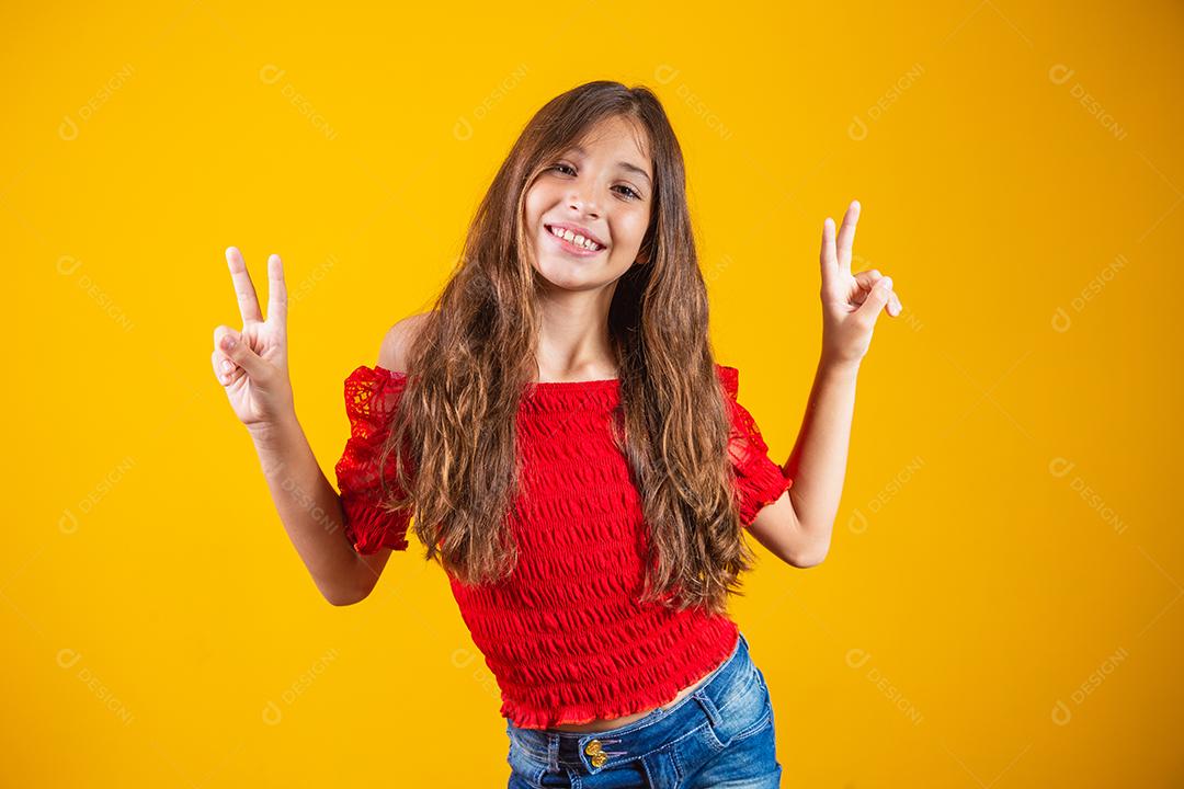 Menina pré-adolescente encantadora e delicada fazendo um sinal de V sorrindo isolado em fundo amarelo.