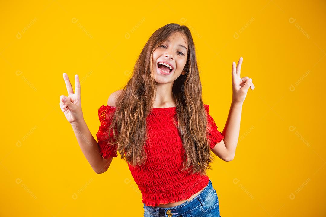 Menina pré-adolescente encantadora e delicada fazendo um sinal de V sorrindo isolado em fundo amarelo.