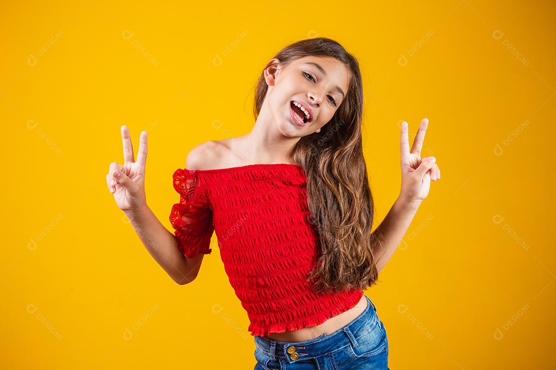 Menina pré-adolescente encantadora e delicada fazendo um sinal de V sorrindo isolado em fundo amarelo.