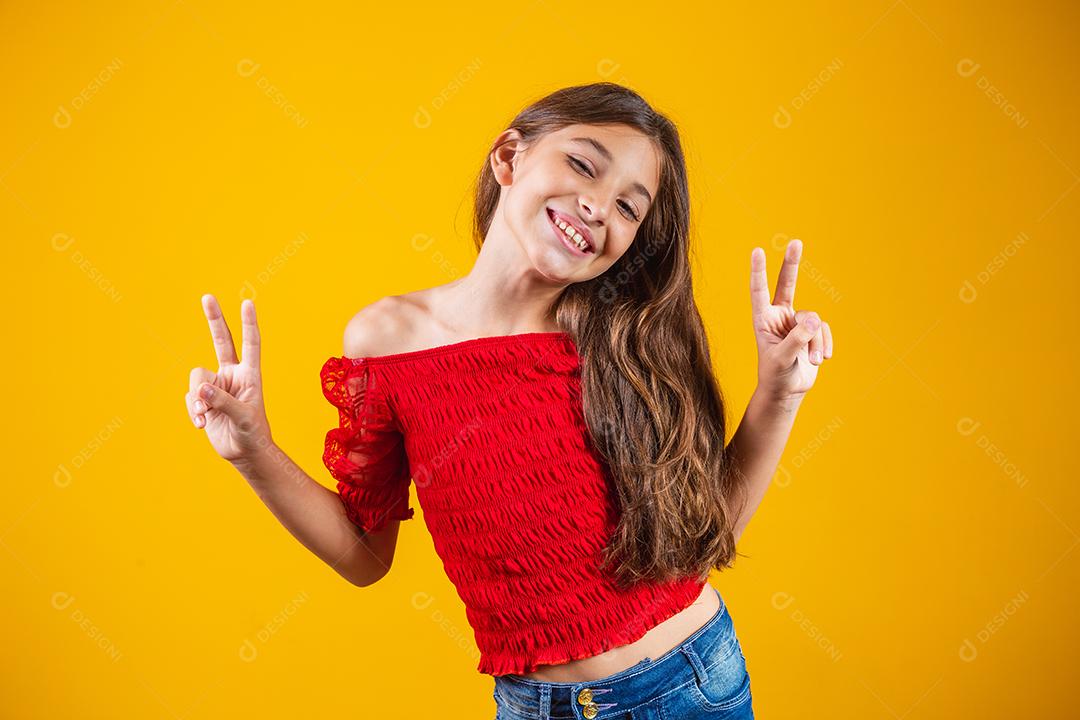 Menina pré-adolescente encantadora e delicada fazendo um sinal de V sorrindo isolado em fundo amarelo.