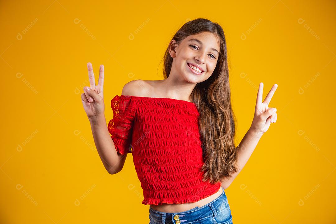 Menina pré-adolescente encantadora e delicada fazendo um sinal de V sorrindo isolado em fundo amarelo.