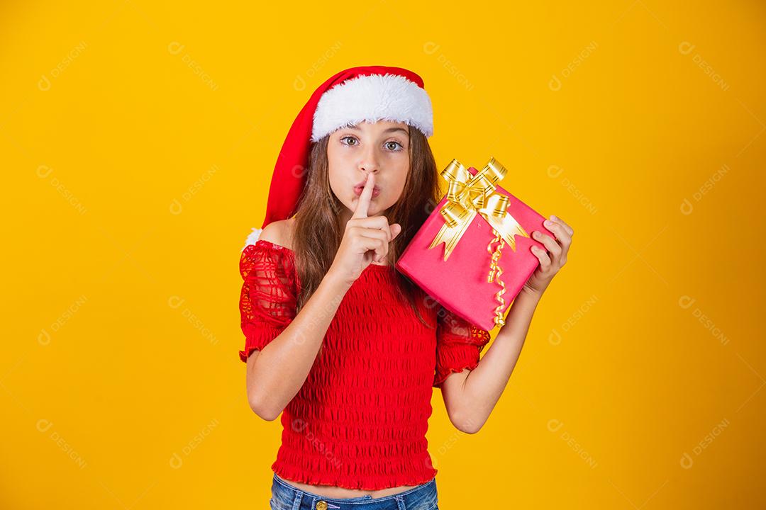pequena garota caucasiana segurando um presente de natal pedindo silêncio à câmera com o dedo na boca