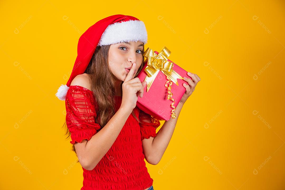 pequena garota caucasiana segurando um presente de natal pedindo silêncio à câmera com o dedo na boca