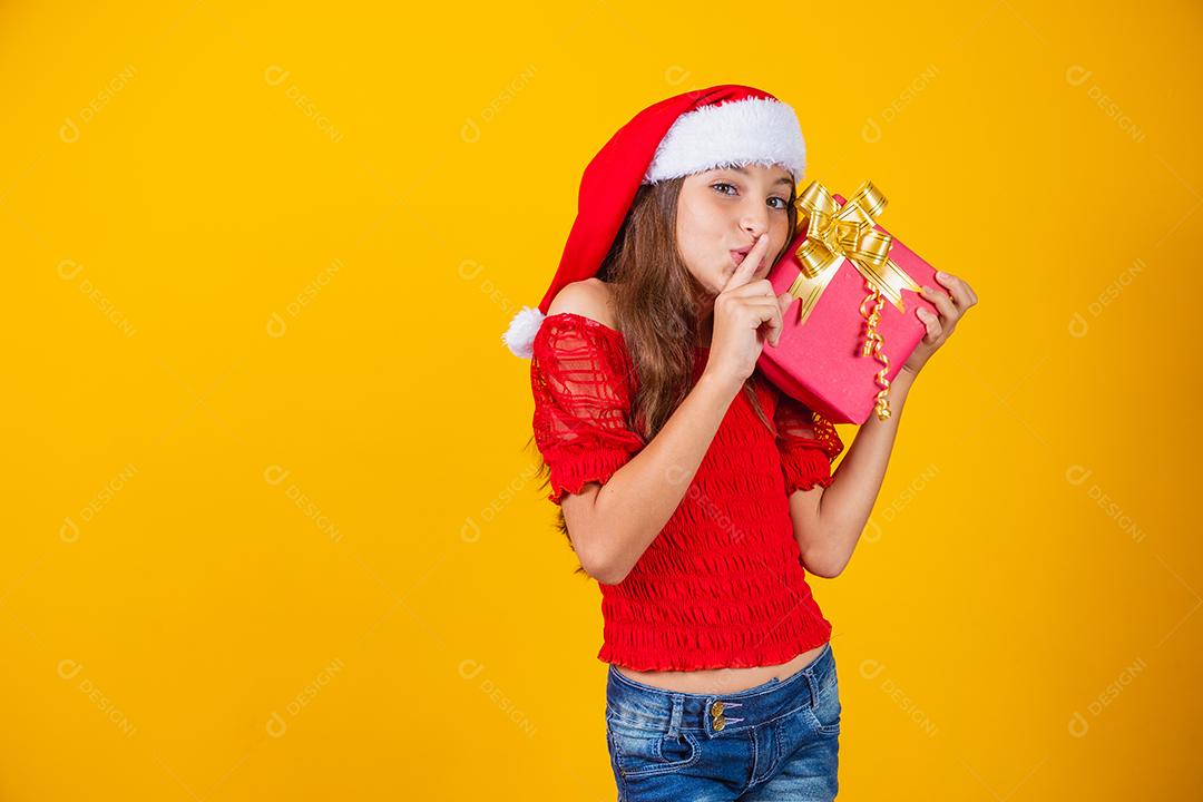 pequena garota caucasiana segurando um presente de natal pedindo silêncio à câmera com o dedo na boca