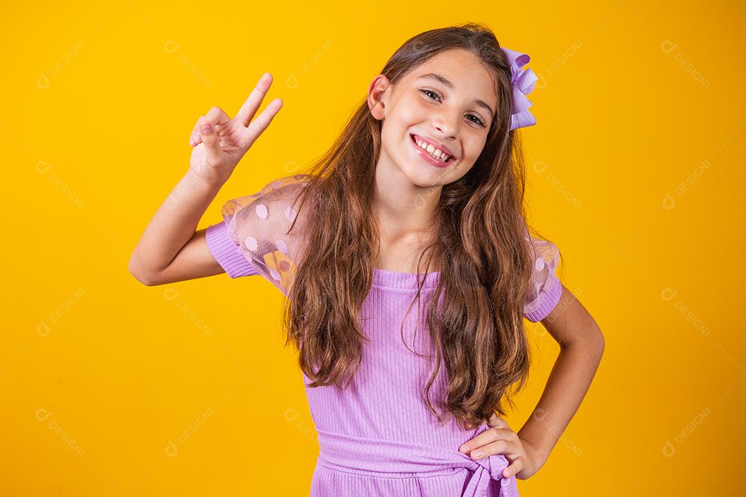 Menina pré-adolescente encantadora e delicada fazendo um sinal de V sorrindo isolado em fundo amarelo.