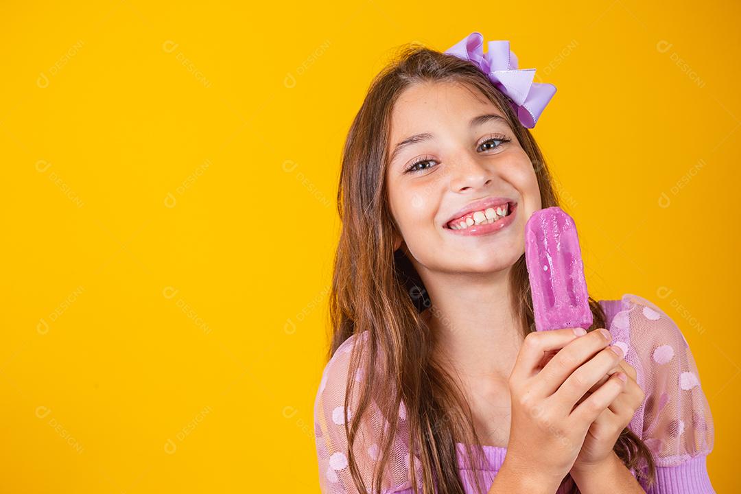 pequena garota caucasiana com delicioso picolé de uva sorrindo olhando para a câmera. Verão