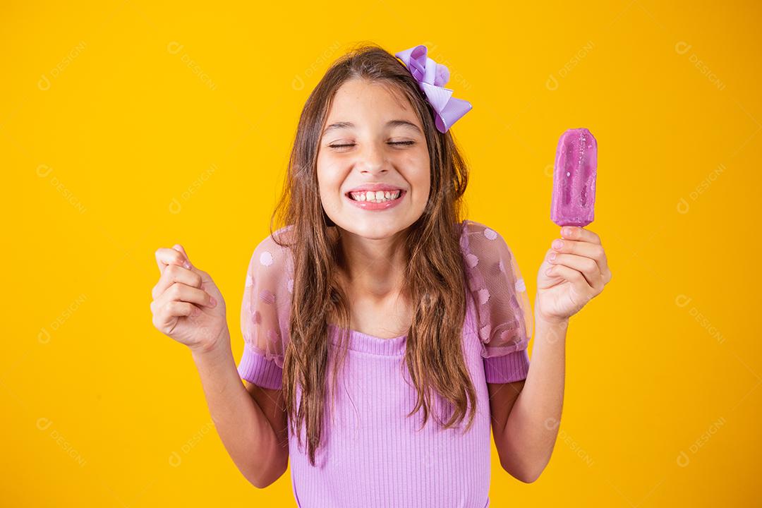 Menina feliz de pele clara come sorvete roxo aromatizado com uvas, cerra o punho com a expectativa
