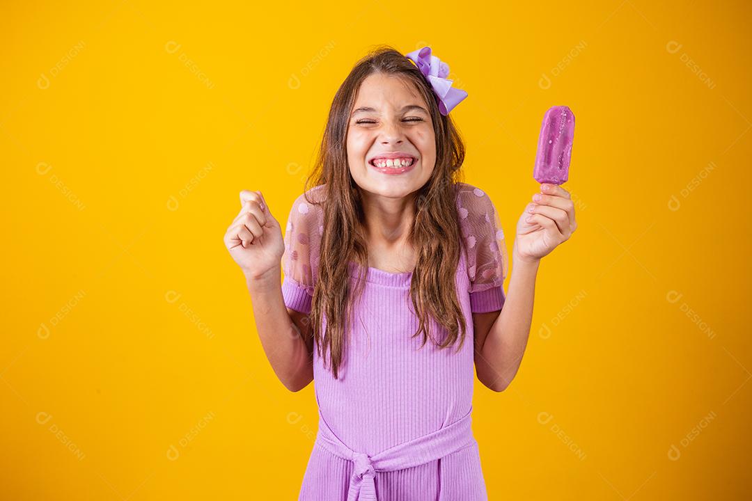 Menina feliz de pele clara come sorvete roxo aromatizado com uvas, cerra o punho com a expectativa