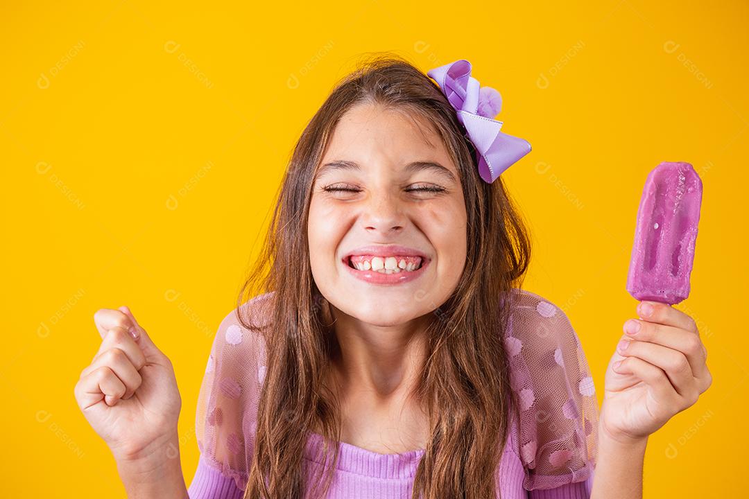 Menina feliz de pele clara come sorvete roxo aromatizado com uvas, cerra o punho com a expectativa
