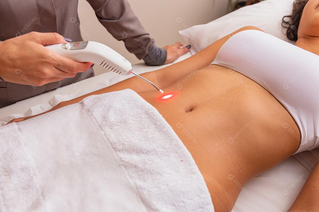 Massagem relaxante e massagem modeladora, drenagem linfática, procedimentos artesanais e estéticosMulher fazendo aplicação contínua e direta de laser terapêutico na artéria radial na barriga no spa. Mulher em tratamento estético com Ilib