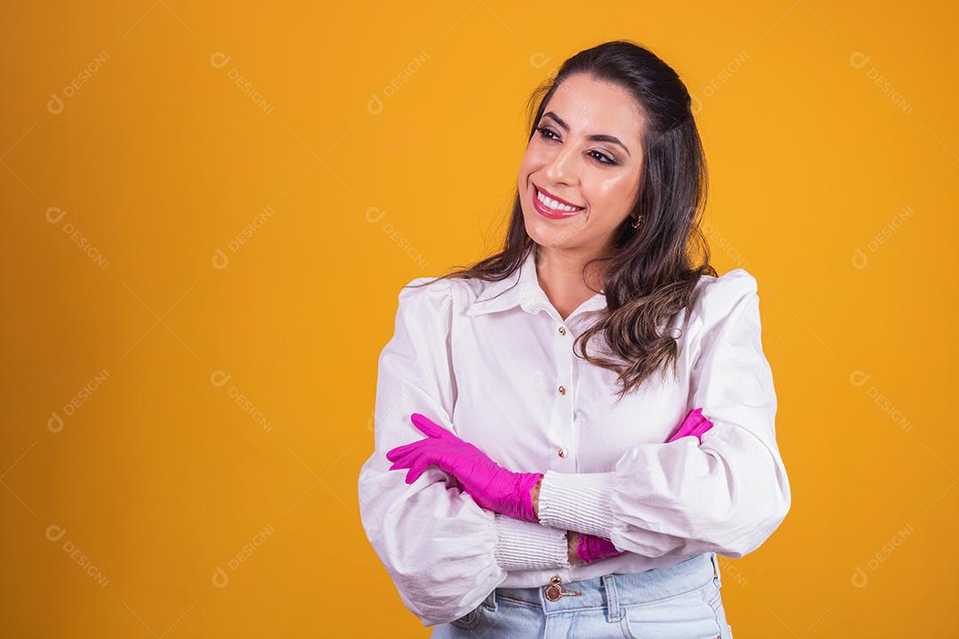 Jovem morena com os braços cruzados sobre fundo amarelo, usando luva rosa.