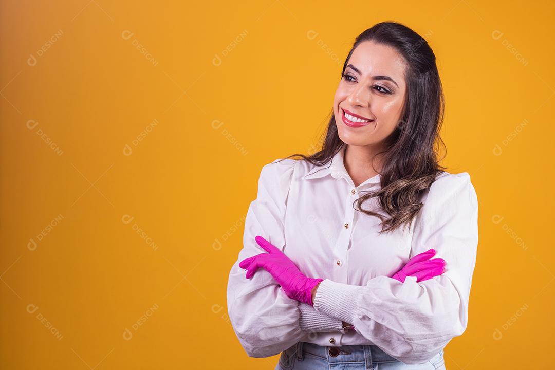Jovem morena com os braços cruzados sobre fundo amarelo, usando luva rosa.
