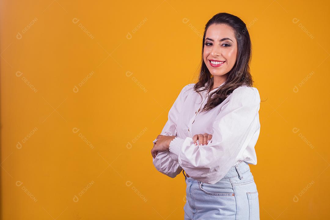 jovem morena com os braços cruzados, olhando para a câmera no fundo amarelo, com espaço livre para texto. Horizontal