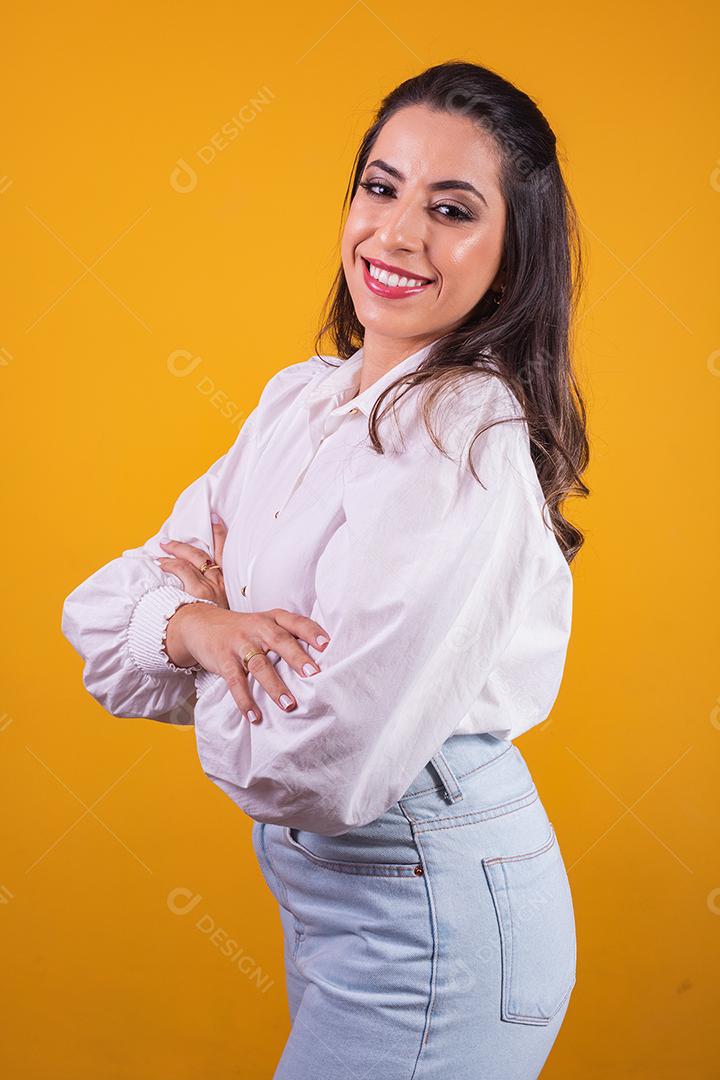 jovem morena com os braços cruzados, olhando para a câmera no fundo amarelo, com espaço livre para texto. Horizontal