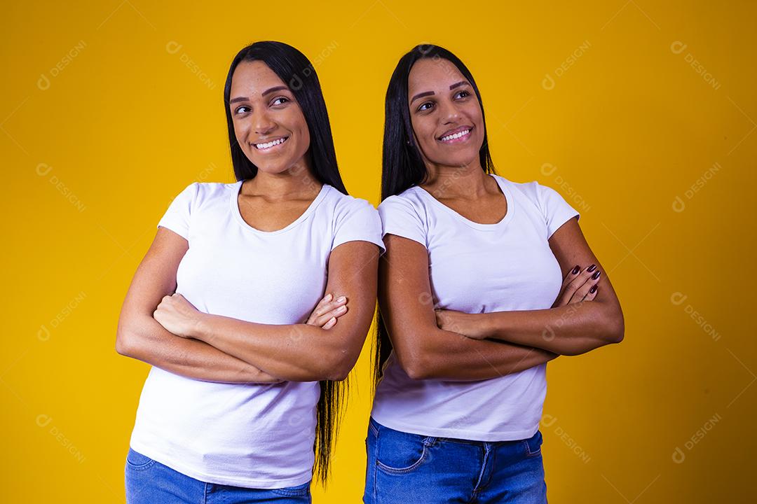 irmãs gêmeas idênticas em fundo amarelo com os braços cruzados