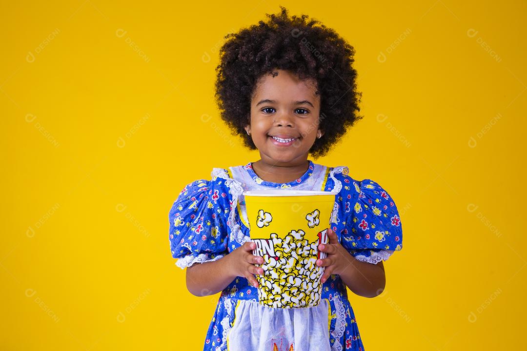 Menina afro bonitinha com roupa de festa junina comendo pipoca em fundo amarelo