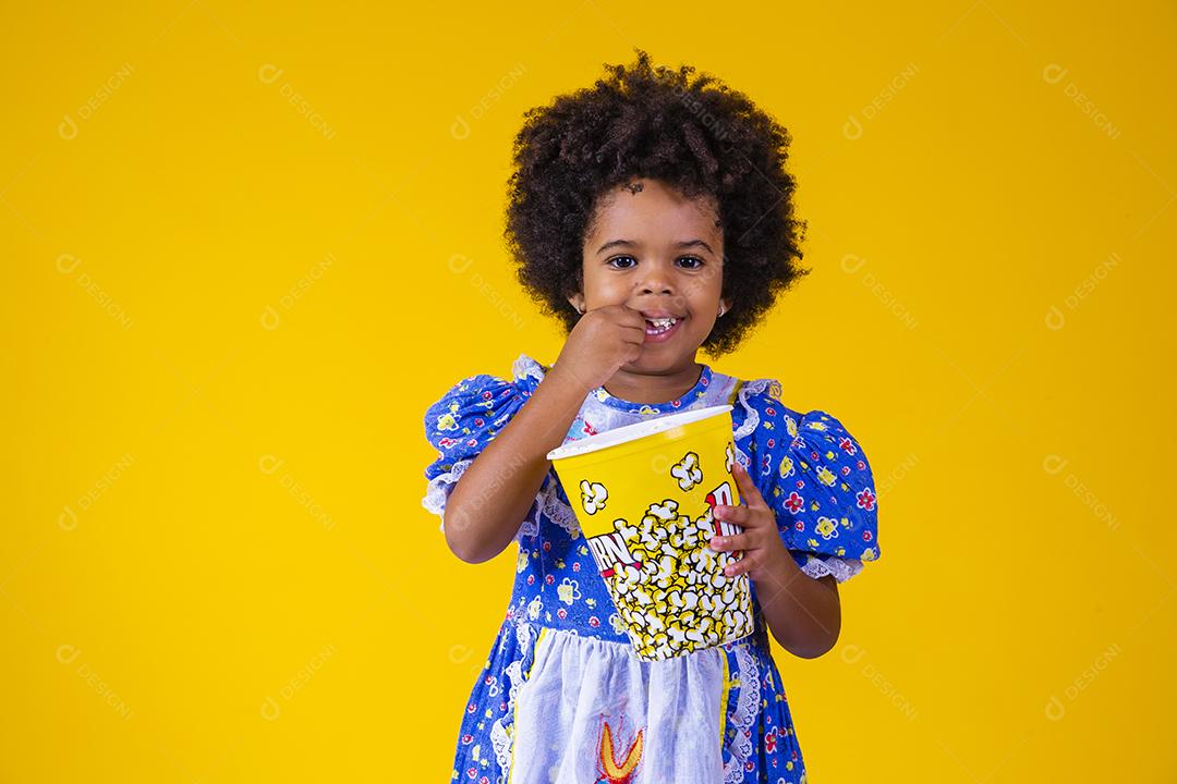 Menina afro bonitinha com roupa de festa junina comendo pipoca em fundo amarelo