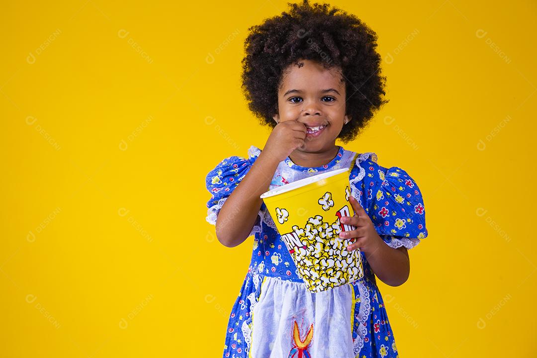 Menina afro bonitinha com roupa de festa junina comendo pipoca em fundo amarelo