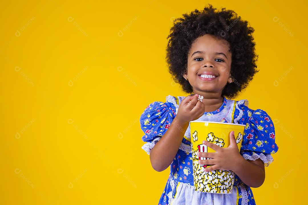 Menina afro bonitinha com roupa de festa junina comendo pipoca em fundo amarelo