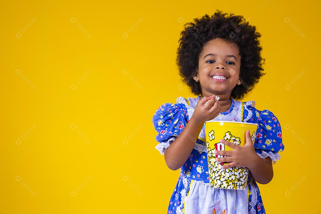 Menina afro bonitinha com roupa de festa junina comendo pipoca em fundo amarelo