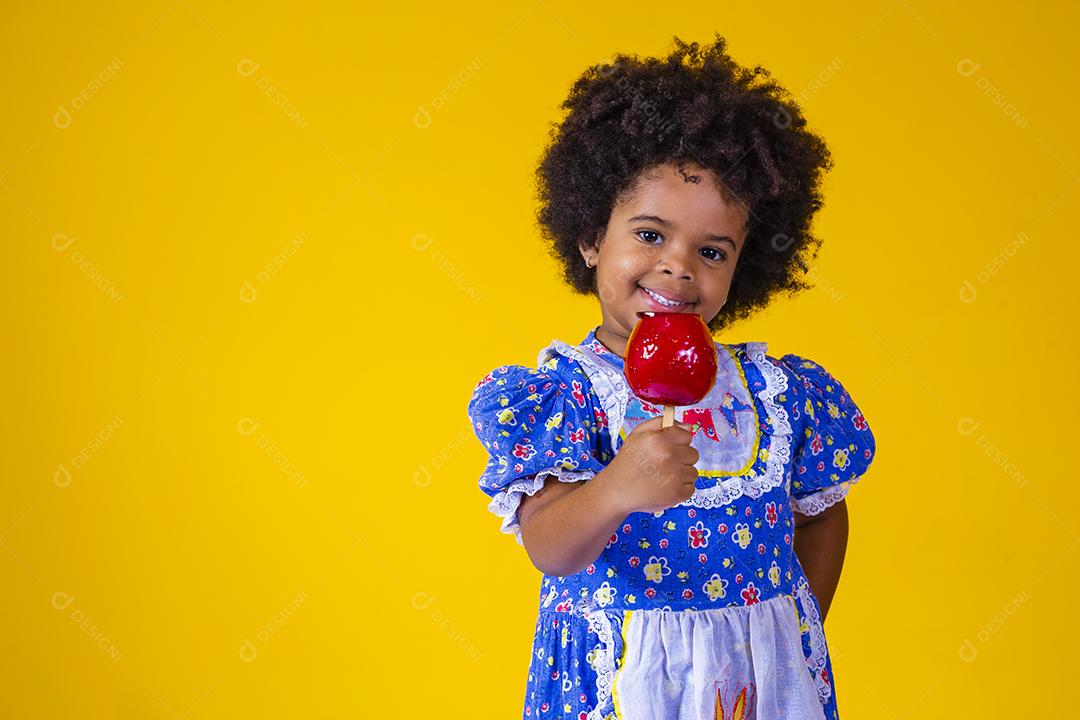 Adorável menina negra vestida com roupas de festa de junho comendo a maçã do amor. Sobremesa de maçã
