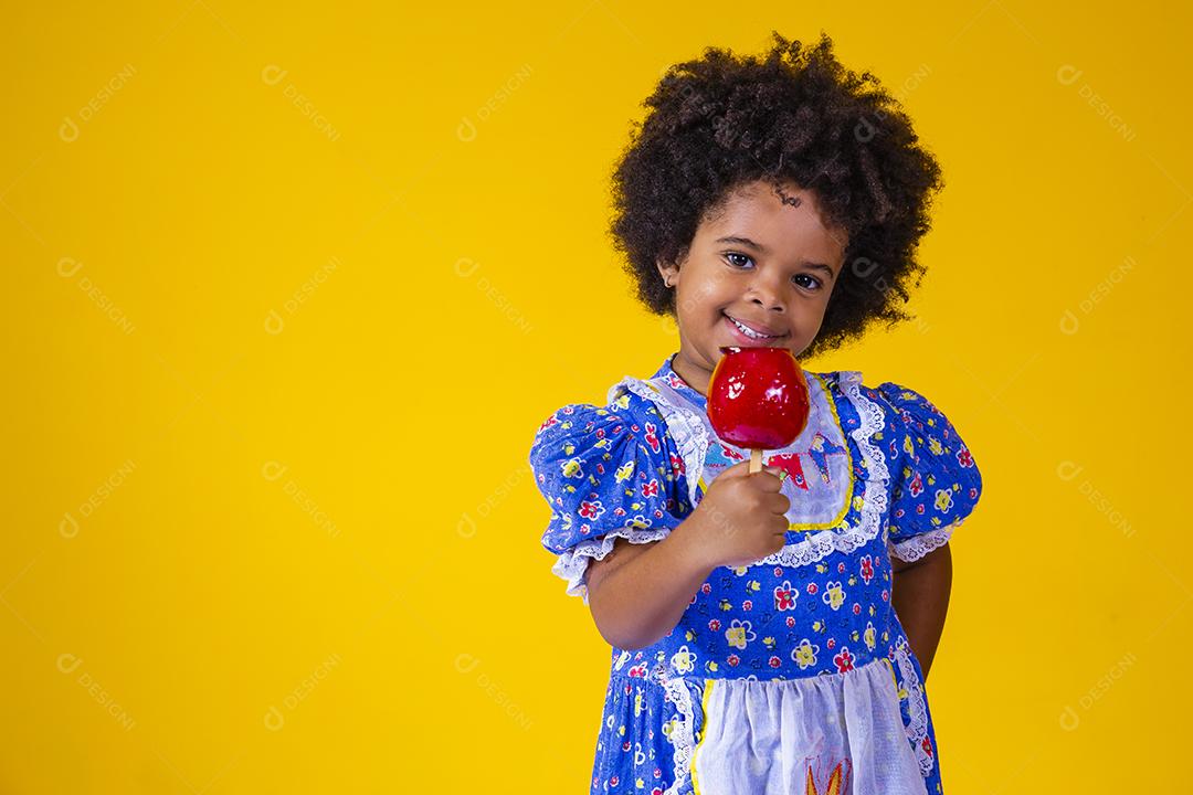 Adorável menina negra vestida com roupas de festa de junho comendo a maçã do amor. Sobremesa de maçã