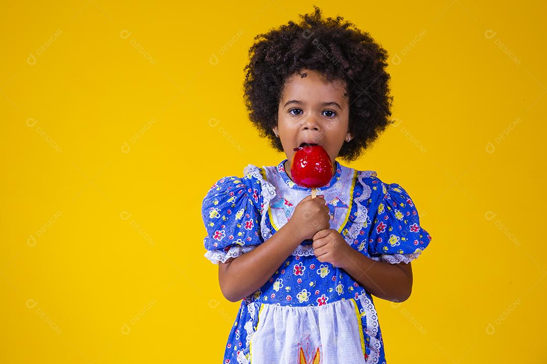 Adorável menina negra vestida com roupas de festa de junho comendo a maçã do amor. Sobremesa de maçã